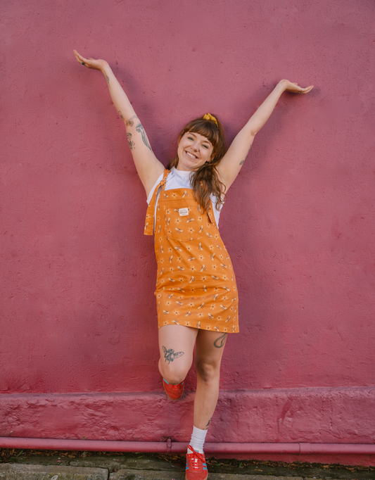 Person wearing an mustard, yellow dungaree/overall dress with a bee pattern against a pink wall