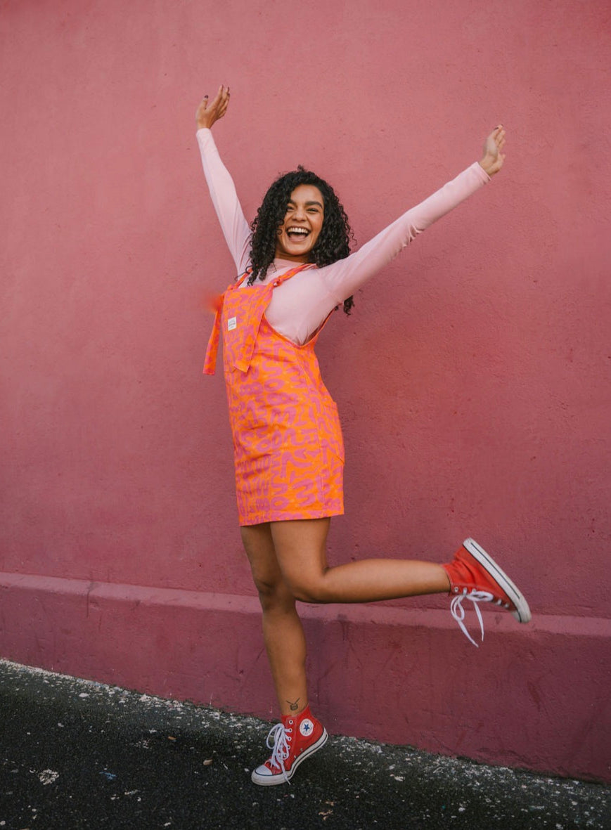 Woman wearing a orange Lamp pattern dungaree/overall/ romper dress standing against a pink wall.