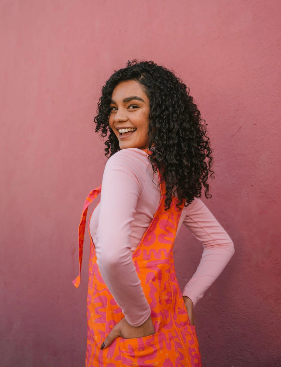 Woman wearing a orange Lamp pattern dungaree/overall/ romper dress standing against a pink wall.