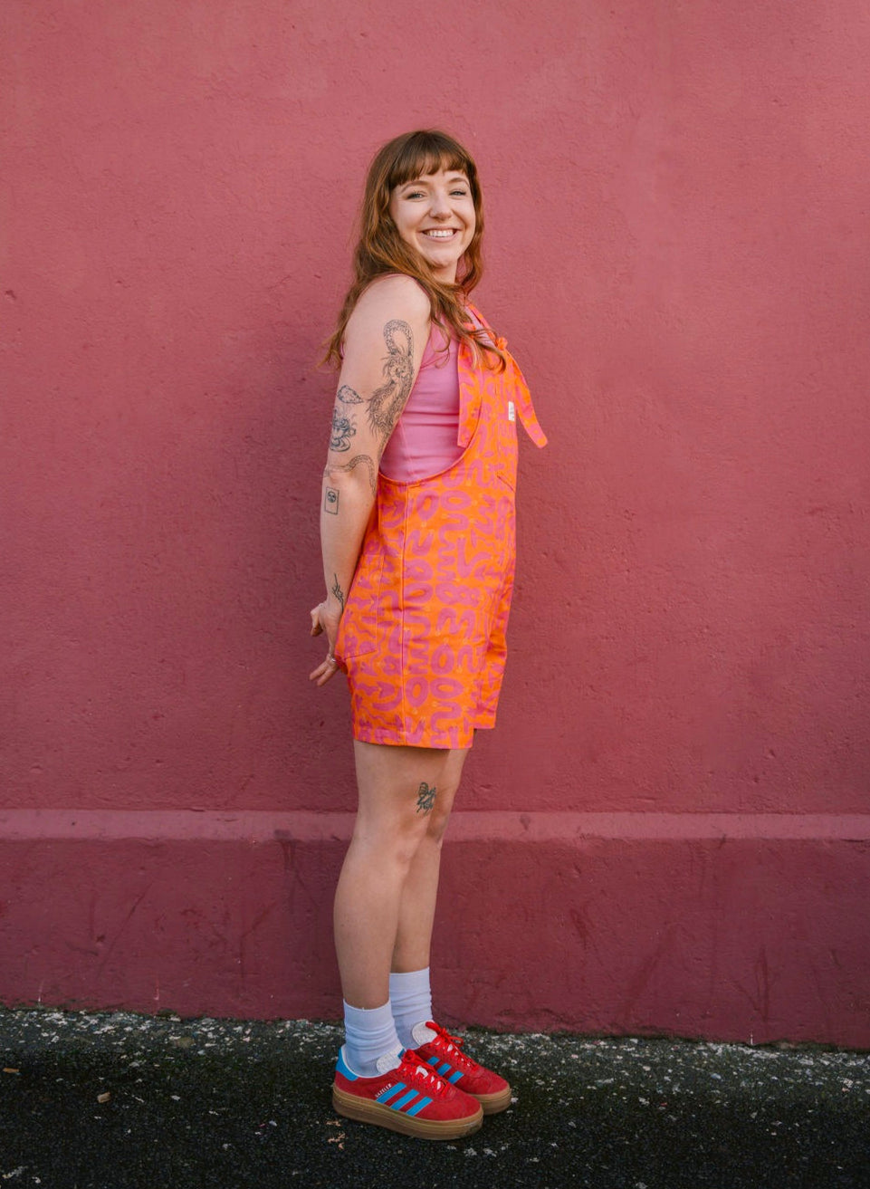 Person wearing a short orange Lamp patterned dungaree/overall/romper standing against a red wall.