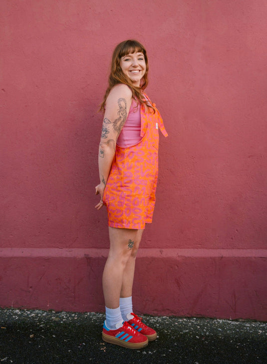 Person wearing a short orange Lamp patterned dungaree/overall/romper standing against a red wall.