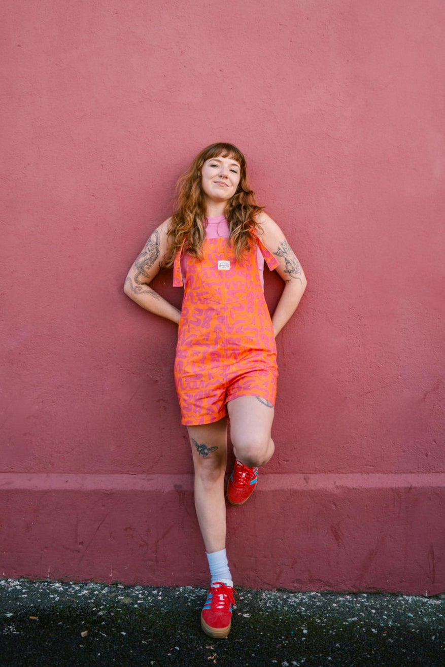 Person wearing a short orange Lamp patterned dungaree/overall/romper standing against a red wall.