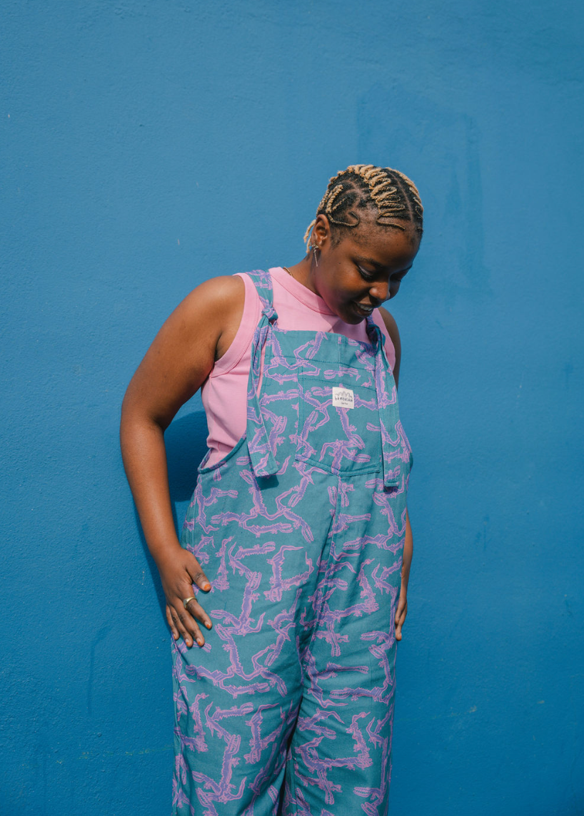 Person wearing a blue, purple patterned dungarees/overalls size Large with a pink crop top against a blue background
