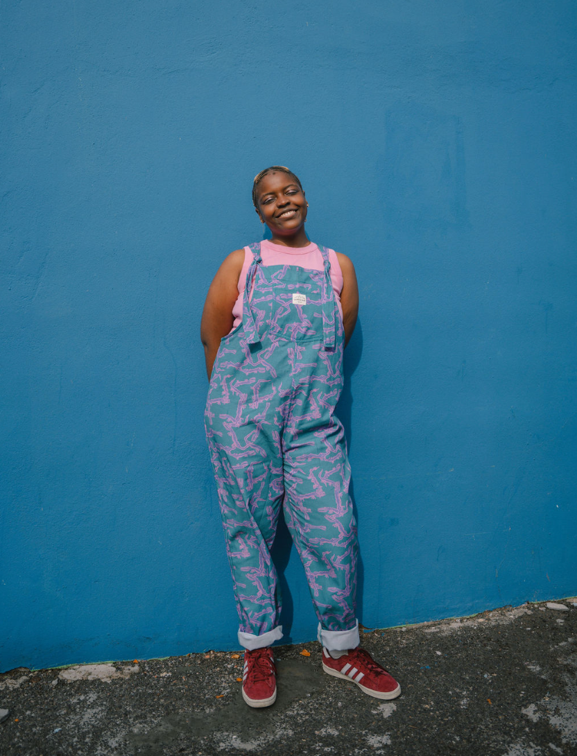 Person wearing a blue, purple patterned dungarees/overalls size Large with a pink crop top against a blue background