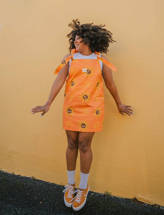 Person wearing an orange sunflower dungaree dress with flower patterns against a yellow wall