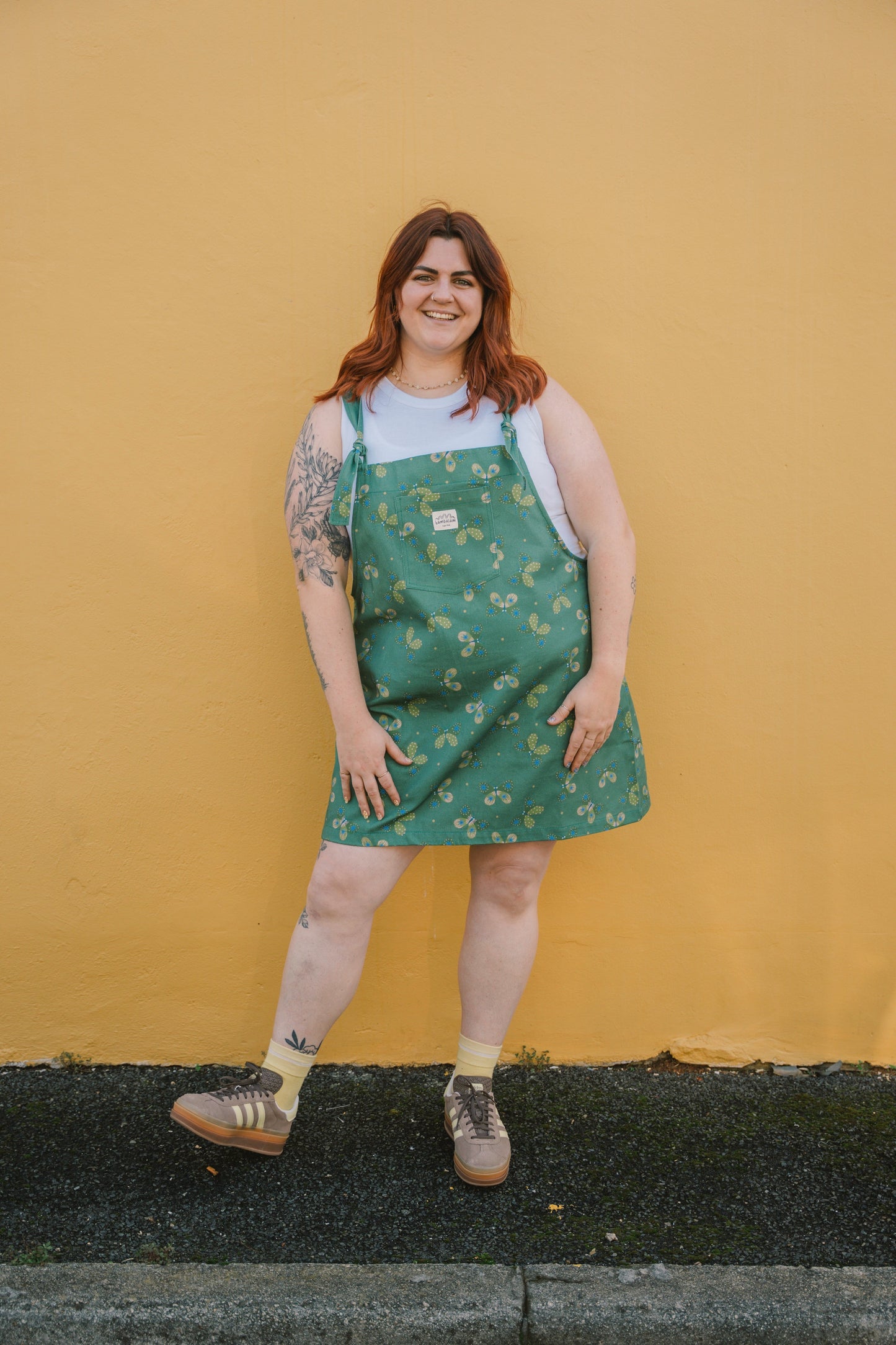 Person wearing a XXL green butterfly dungaree dress with a pattern against a yellow wall.