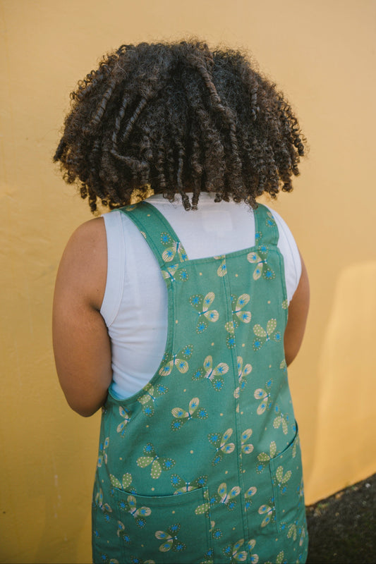 Person wearing a green butterfly patterned dungaree dress with a yellow wall in the background