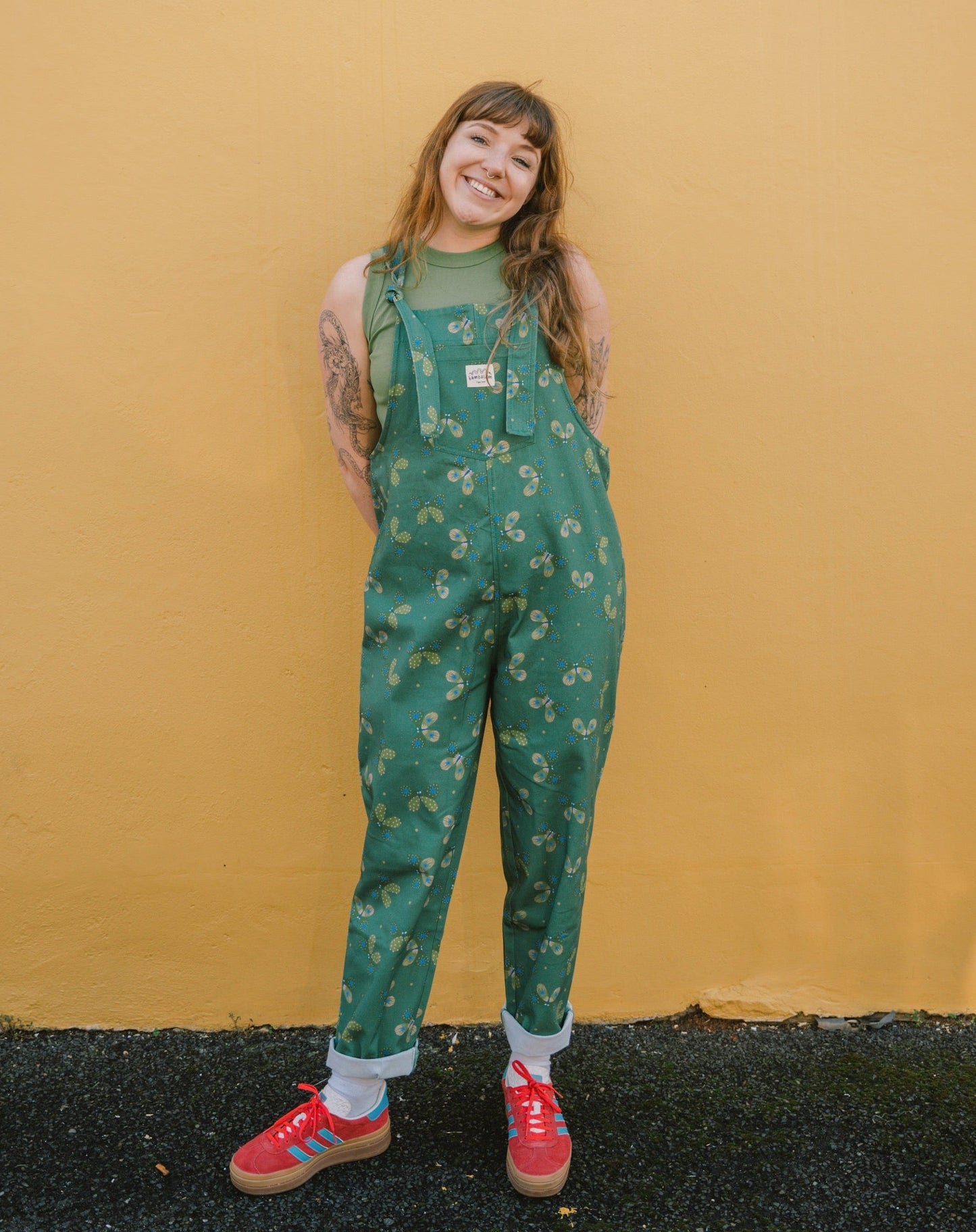 Person wearing green Butterfly long patterned overalls/dungarees against a yellow wall