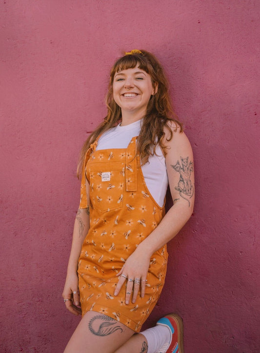 Girl wearing an mustard, yellow dungaree/overall dress with a bee pattern against a pink wall