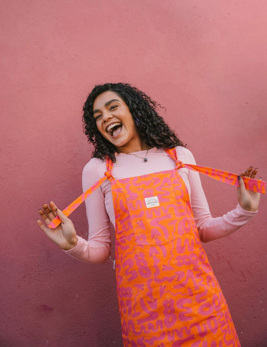 Woman wearing a orange Lamp pattern dungaree/overall/ romper dress standing against a pink wall.