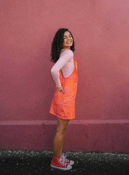 Woman wearing a orange Lamp pattern dungaree/overall/ romper dress standing against a pink wall.