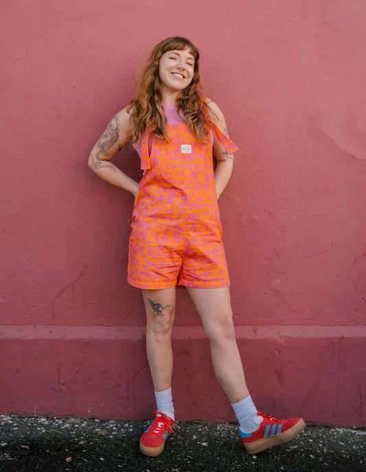 Person wearing a short orange Lamp patterned dungaree/overall/romper standing against a red wall.