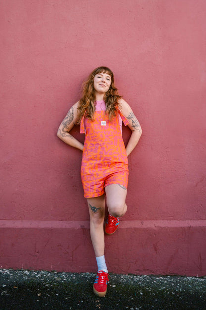 Person wearing a short orange Lamp patterned dungaree/overall/romper standing against a red wall.
