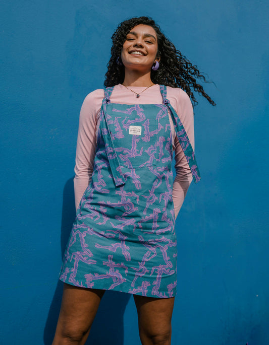 Woman wearing a blue, purple and pink patterned dungaree/overalls dress against a blue background